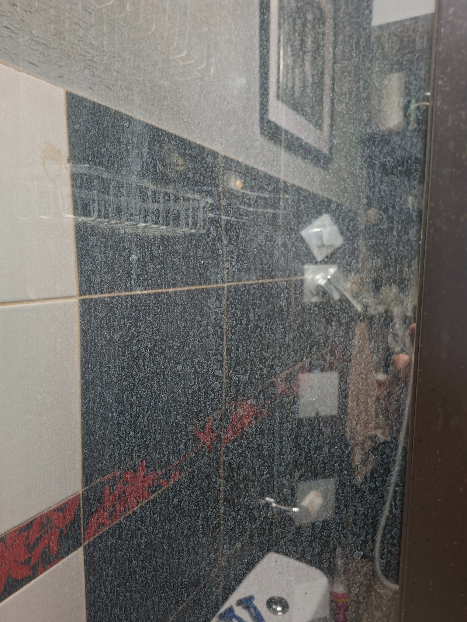 Zaschnięte zacieki na szklanej ściance kabiny prysznicowej, widoczne płytki z dekoracyjnym wzorem i akcesoria łazienkowe. Wymaga dokładnego czyszczenia.