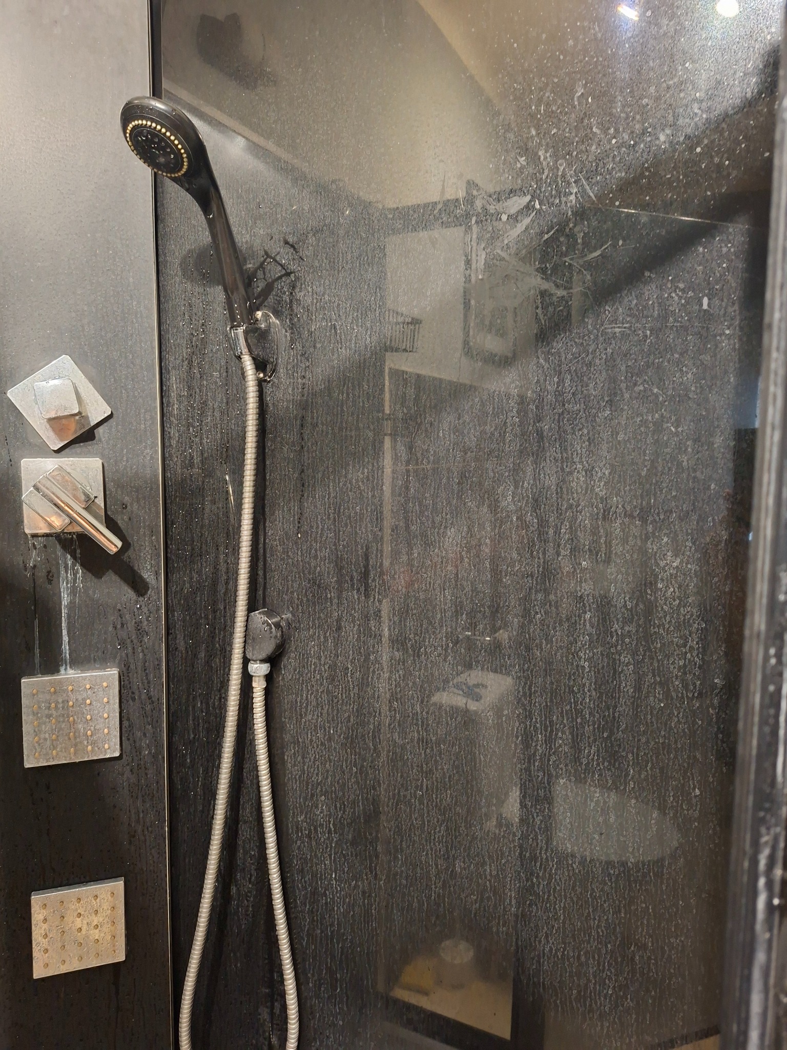 Zabrudzona kabina prysznicowa z czarną armaturą, widoczne zacieki i osad na szklanych drzwiach. Potrzeba gruntownego czyszczenia.