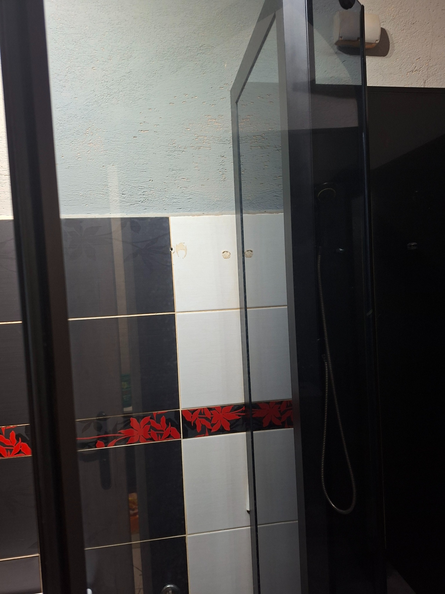 Kabina prysznicowa z czarnymi ramami i wzorzystymi płytkami w łazience. Widoczne ślady użytkowania na fugach i płytkach.