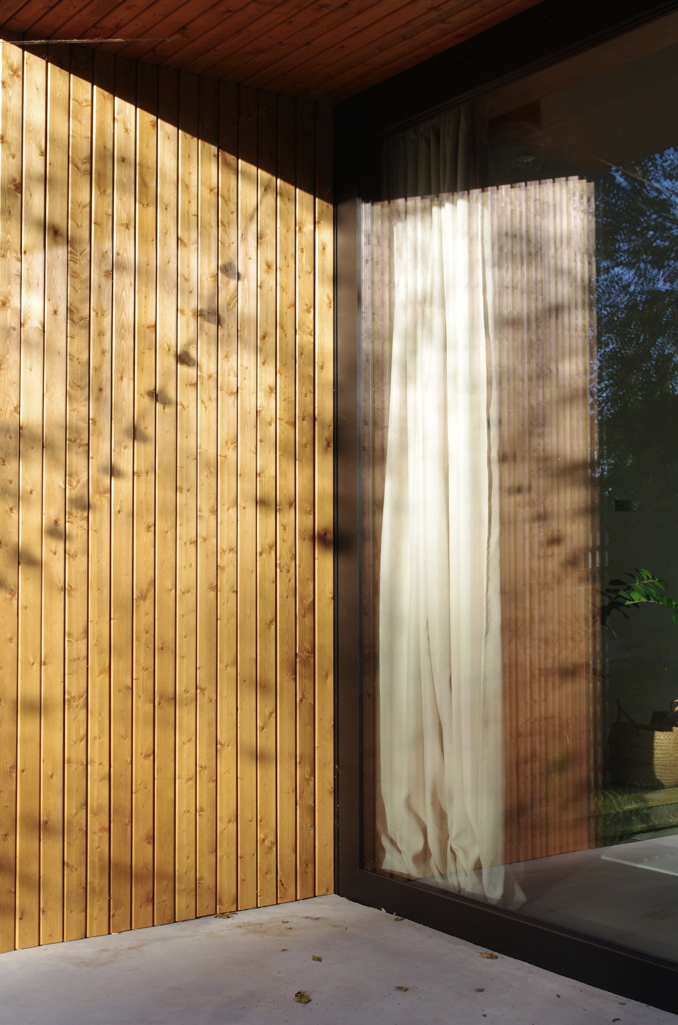 Nowoczesna elewacja domu z drewnianą okładziną i dużym oknem z zasłoną. Widoczne refleksy światła i cienia na drewnie. Minimalistyczny design.