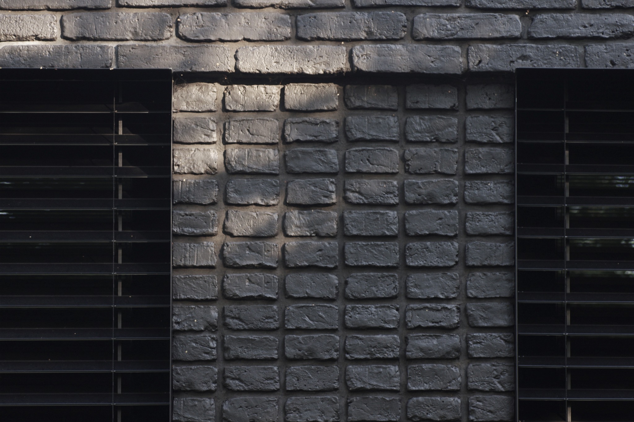 Czarna, ceglana elewacja budynku z żaluzjami okiennymi. Fragment ściany w mocnym kontraście światła i cienia, podkreślającym fakturę cegieł.