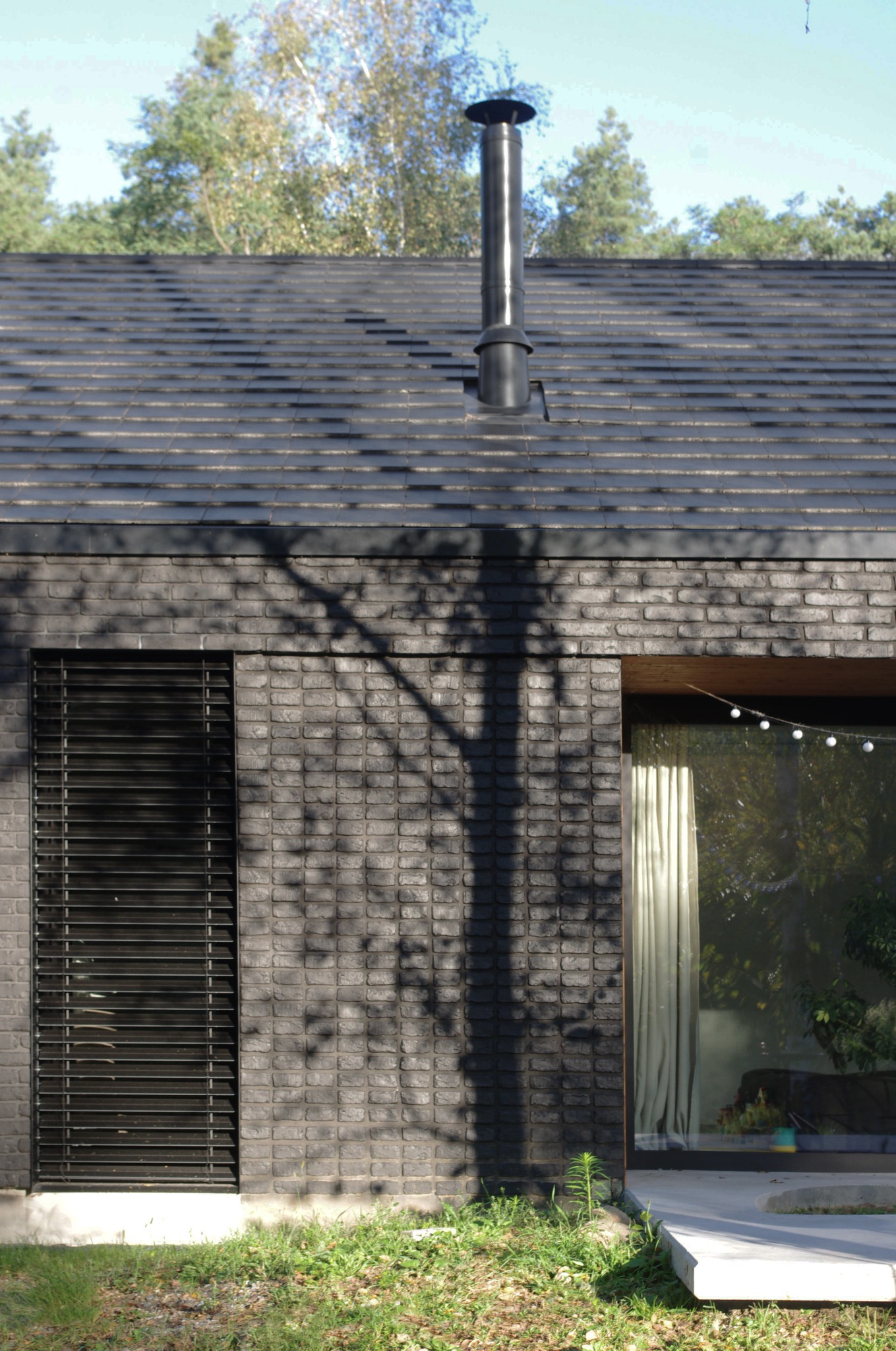 Ciemna elewacja domu z cegły z czarnymi żaluzjami i oknem, na którym widać odbicie drzewa. Na dachu komin. Detal elewacji w nowoczesnym stylu.