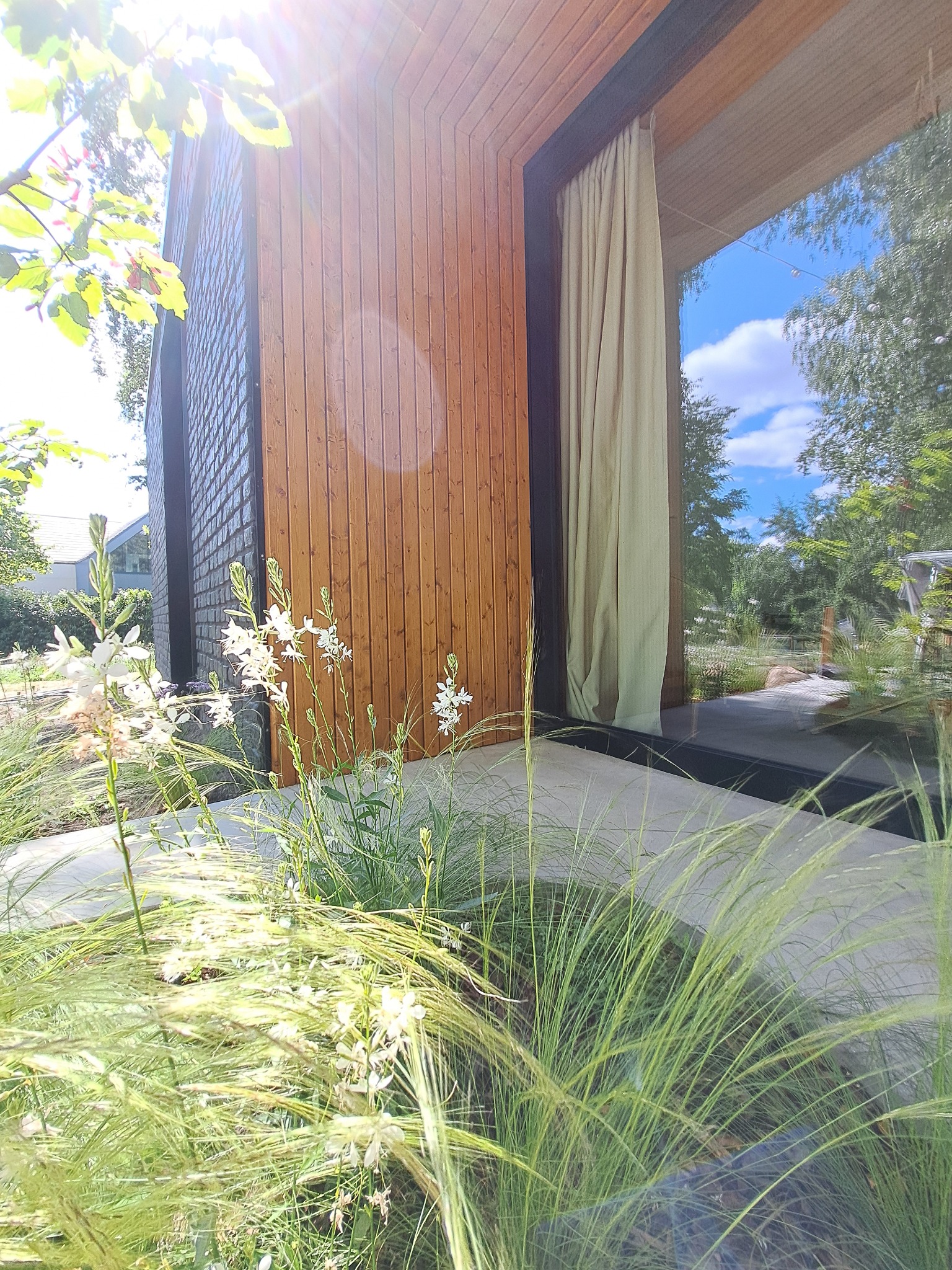 Nowoczesna elewacja domu z drewnianą okładziną, dużym oknem z zasłoną i elementami zieleni w otoczeniu. Widok z poziomu ogrodu.