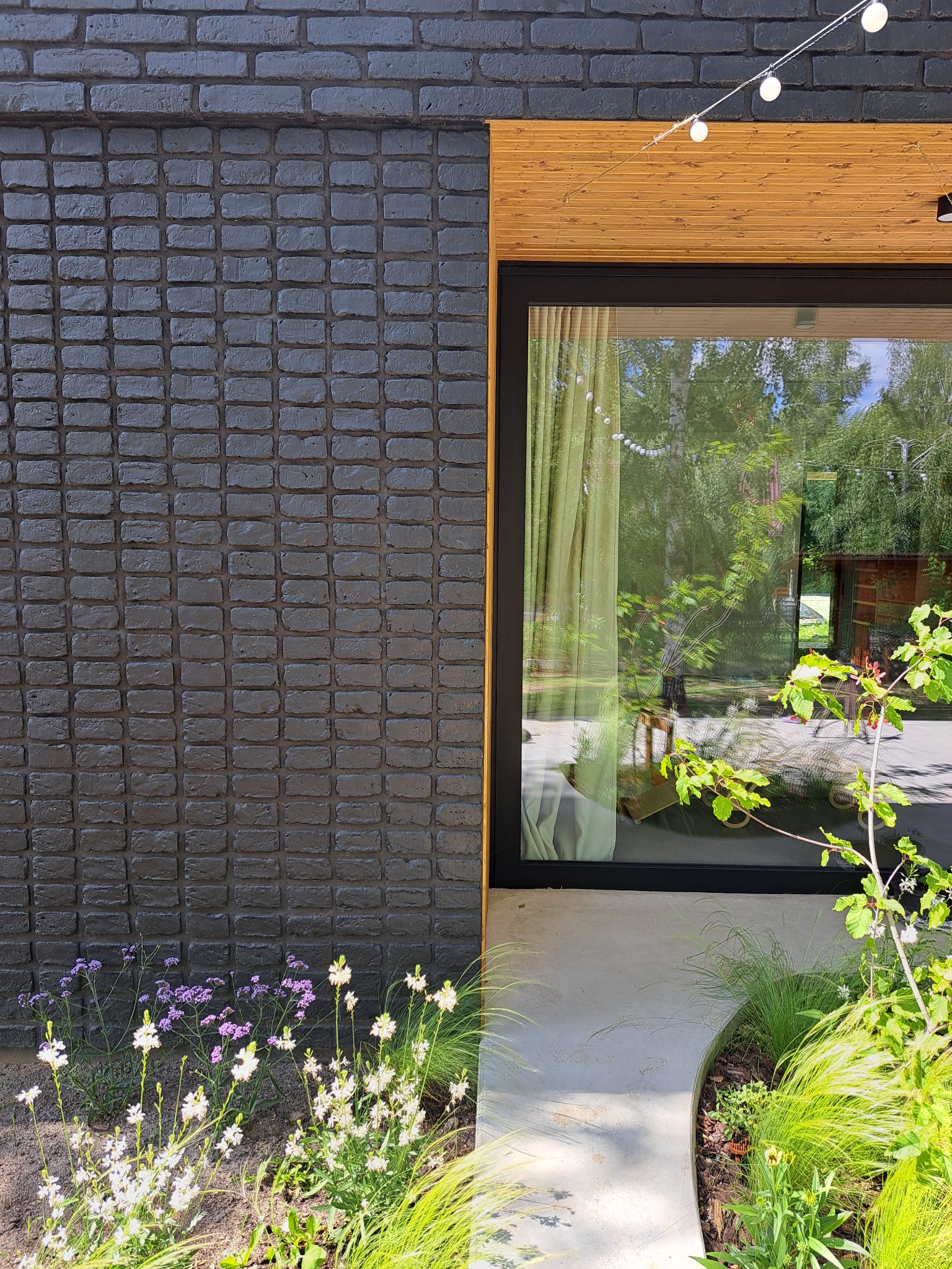 Elewacja z ciemnej cegły i dużego okna z drewnianą ramą. Ogród z kwiatami i trawami. Nowoczesny design. Oświetlenie dekoracyjne.
