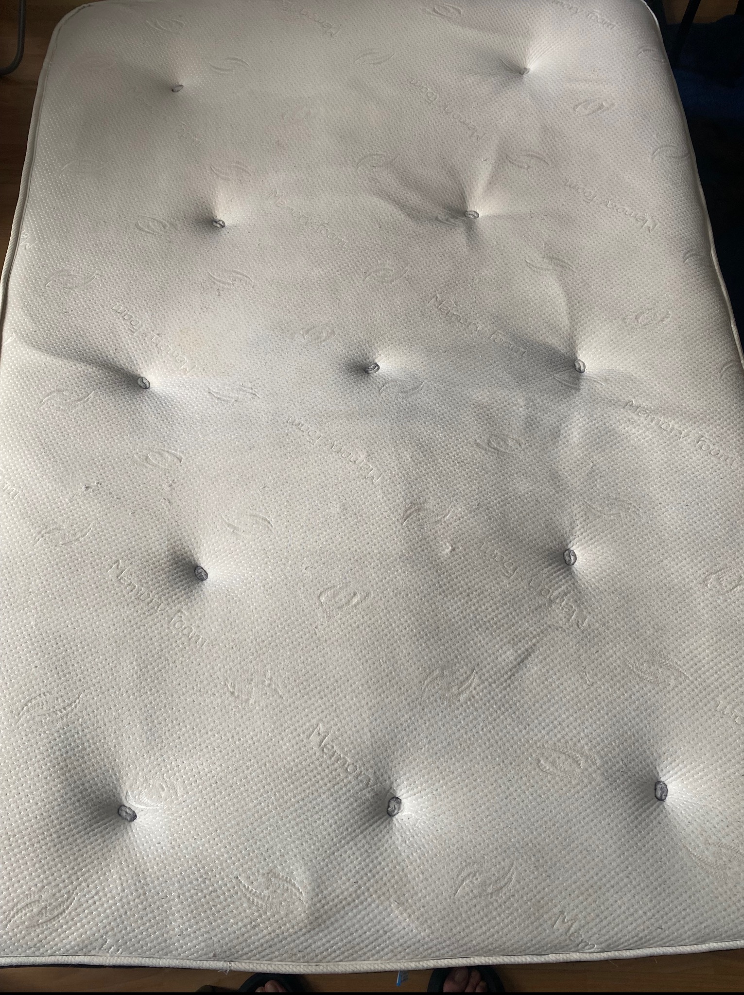 Biały materac z guzikami i wzorem 'Memory Foam', sfotografowany z góry, ukazujący fakturę i detale wykończenia. Widoczne lekkie zabrudzenia.