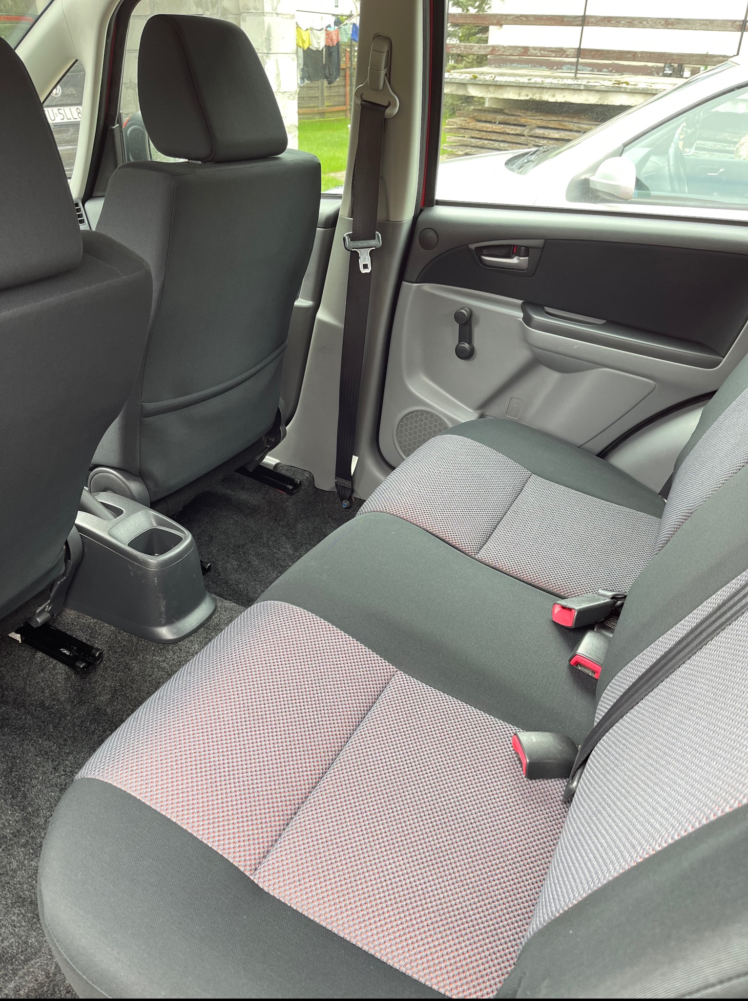 Wnętrze samochodu: tylna kanapa z szarą tapicerką w kratkę, czarne zagłówki i pasy bezpieczeństwa. Widok na podłogę i elementy karoserii.