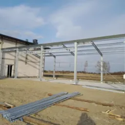 Konstrukcja stalowa hali w trakcie budowy w Stanin, widoczne elementy montażowe na ziemi i budynek w tle.