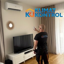 KLIMAT KONTROL Krzysztof Zbyszyński - Instalacja klimatyzacji w salonie: monter z pilotem celuje w biały klimatyzator Haier. Widoczne logo firmy na koszulce montera i grafika 'Klimat Kontrol'.