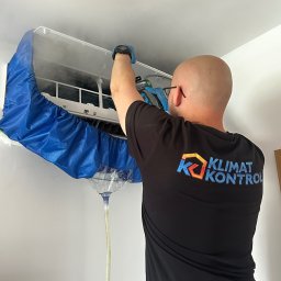 KLIMAT KONTROL Krzysztof Zbyszyński - Serwisant w czarnej koszulce z logo czyści klimatyzator w mieszkaniu, używając specjalnego pokrowca i węża odprowadzającego wodę, co zapewnia czystość podczas serwisu.