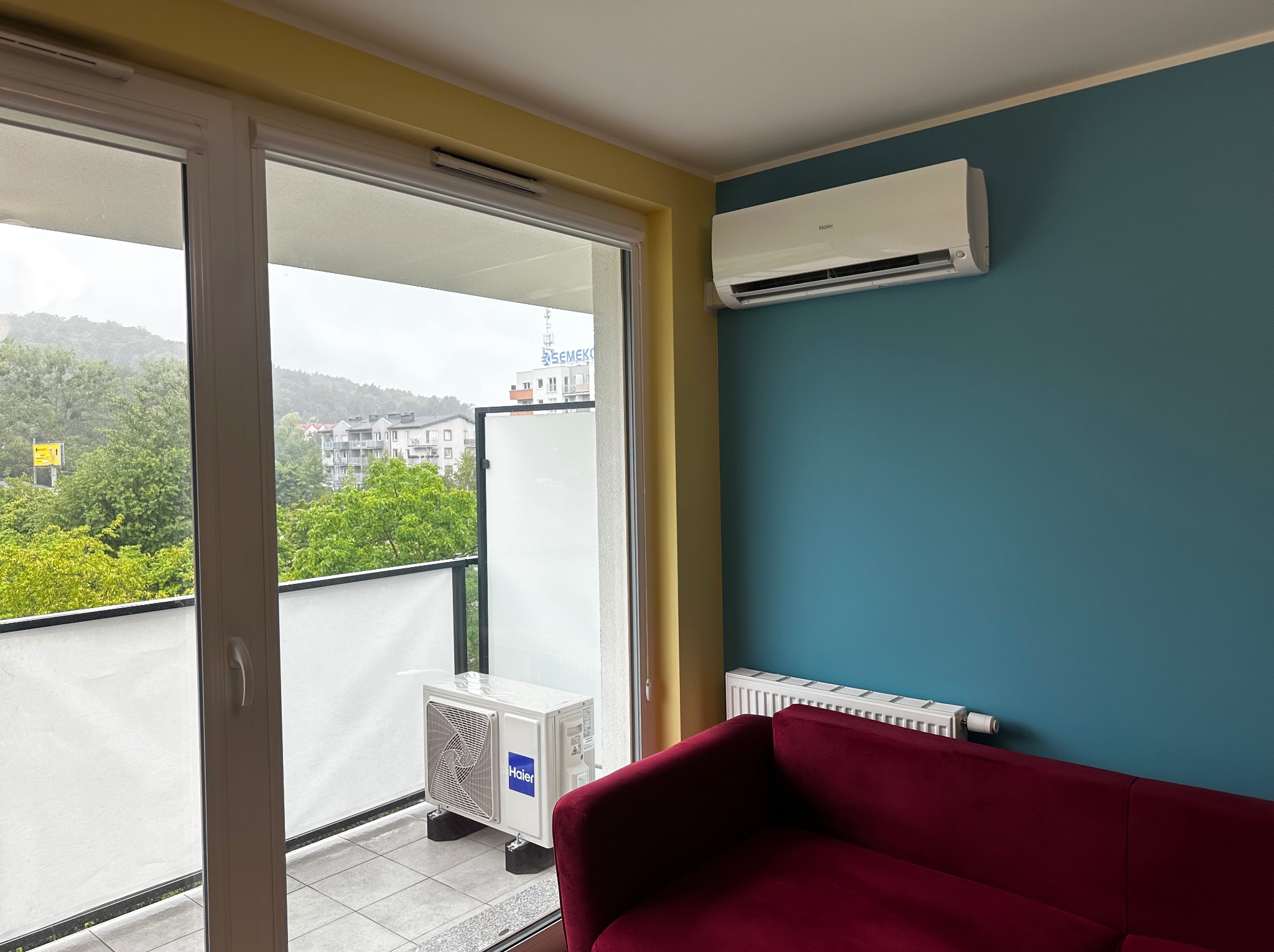 Nowoczesny klimatyzator Haier na ścianie w pokoju z balkonem. Widoczny również grzejnik i fragment czerwonej sofy. Na balkonie jednostka zewnętrzna klimatyzacji.
