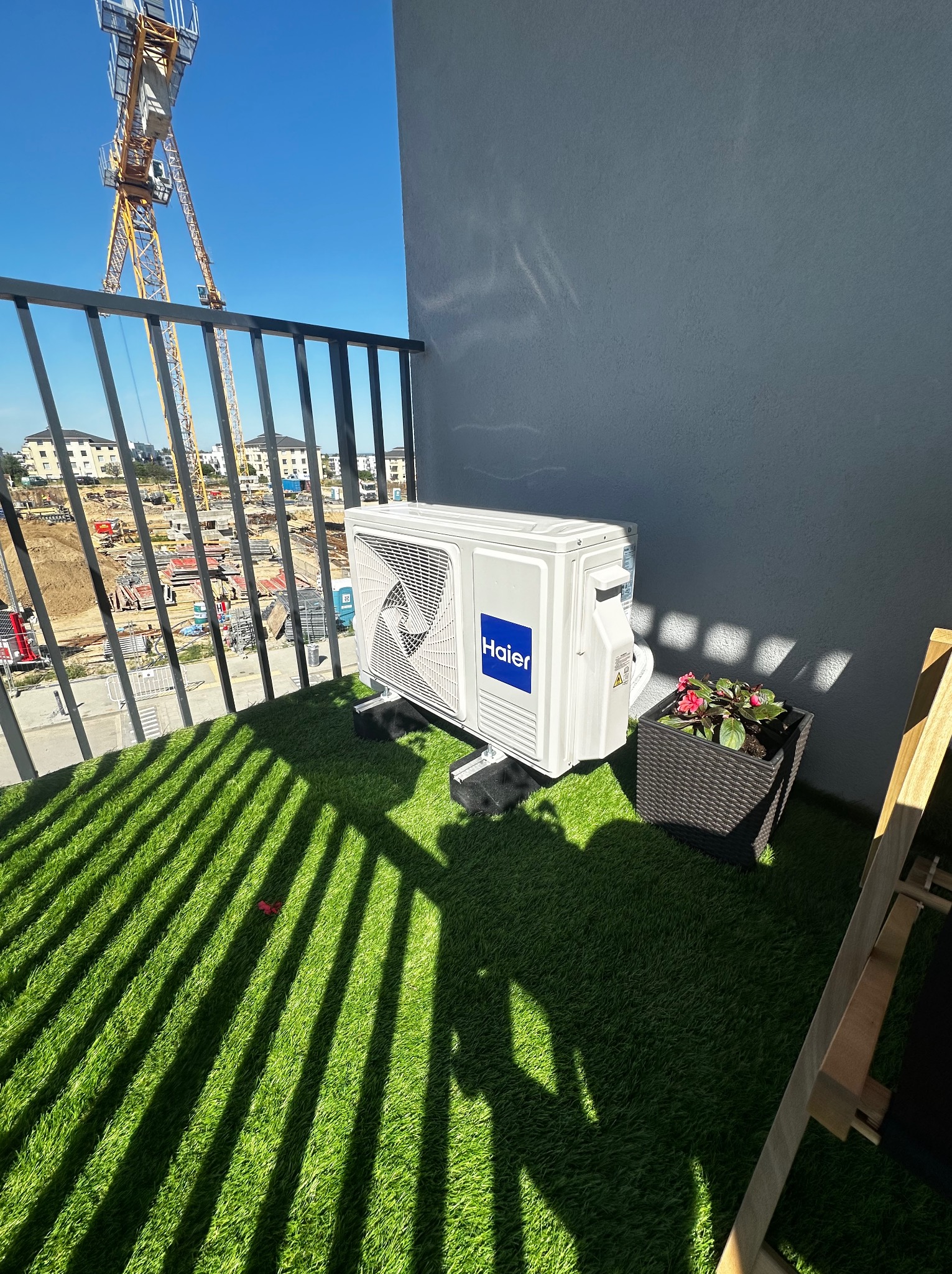 Biała jednostka zewnętrzna klimatyzacji Haier na balkonie z sztuczną trawą, widok na plac budowy i żuraw. Cień barierki na podłożu.