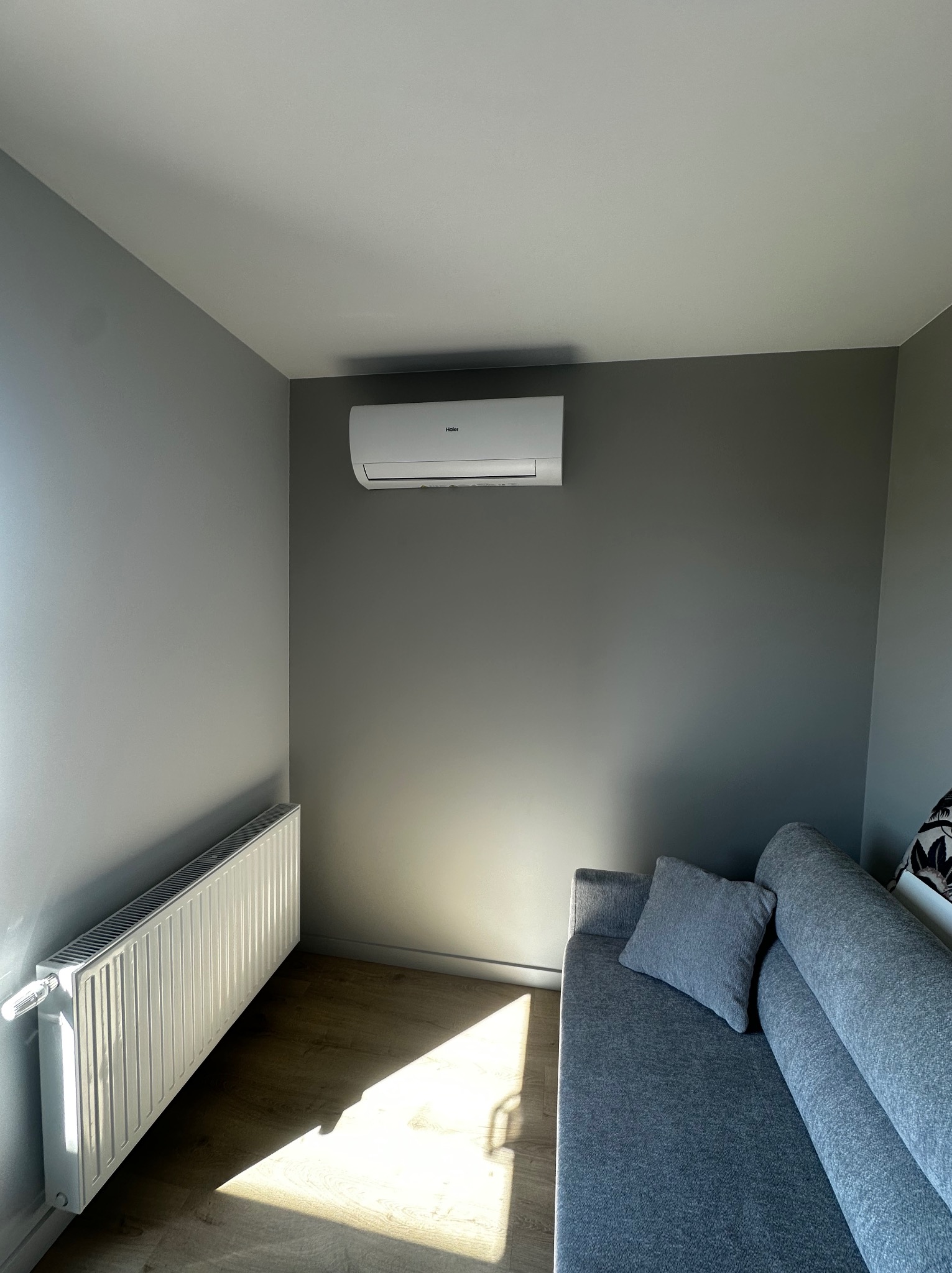 Szary pokój z klimatyzatorem Haier, białym grzejnikiem i szarą sofą. Promienie słoneczne oświetlają podłogę. Minimalistyczny wystrój wnętrza.