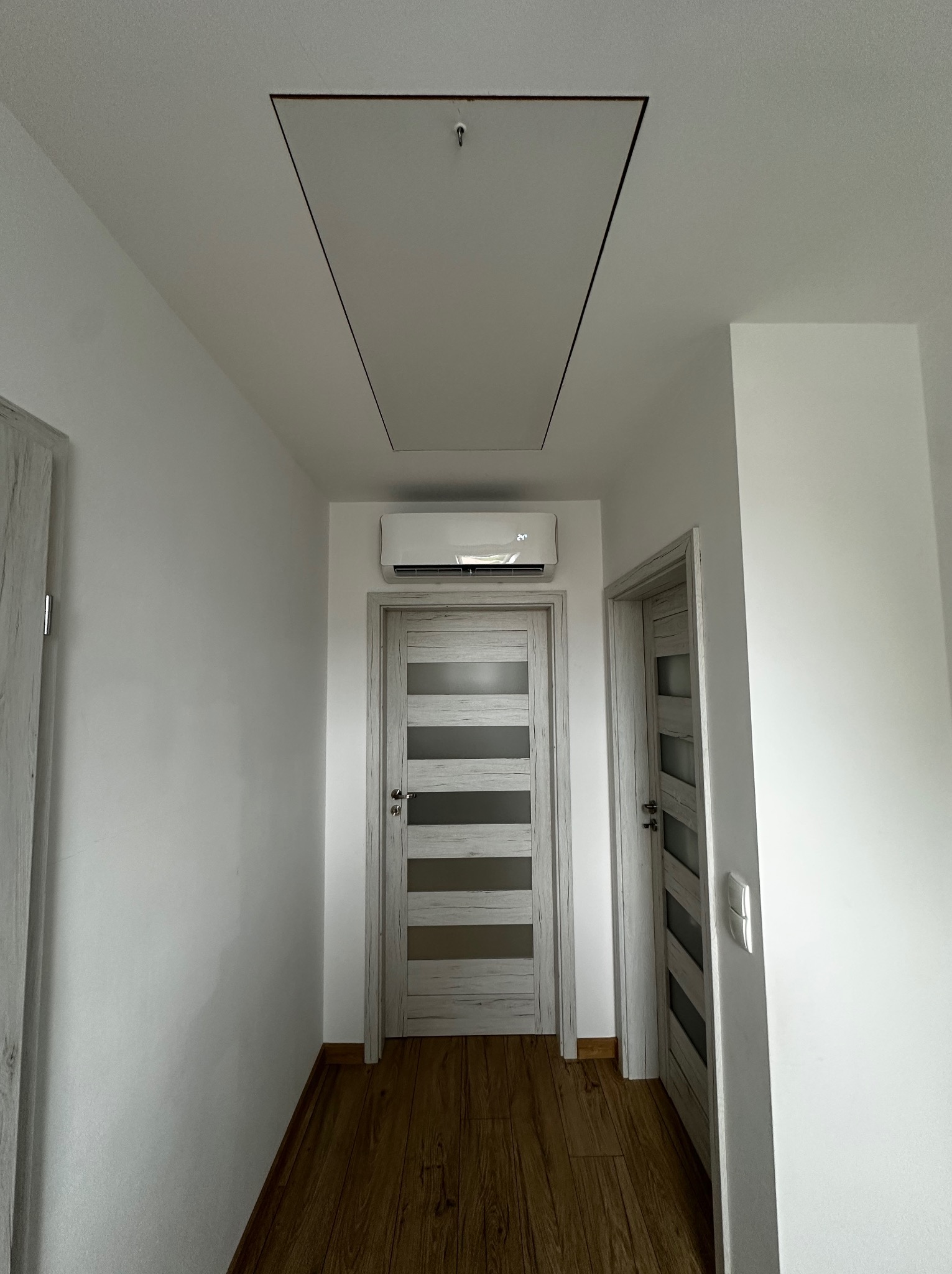 Nowoczesny, biały klimatyzator ścienny zamontowany nad drzwiami w jasnym korytarzu z drewnianą podłogą i klapą rewizyjną w suficie.