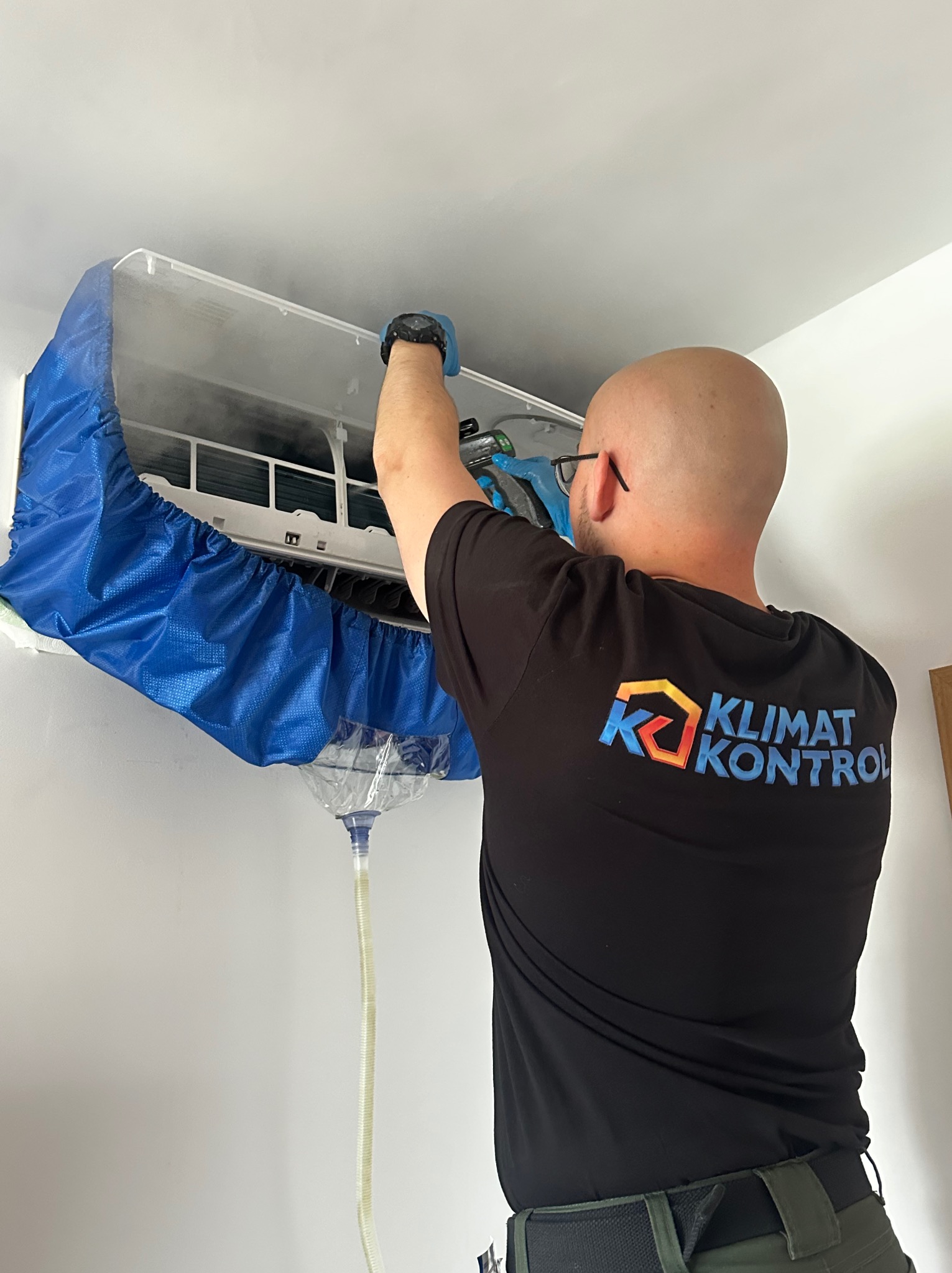 Serwisant w czarnej koszulce z logo czyści klimatyzator w mieszkaniu, używając specjalnego pokrowca i węża odprowadzającego wodę, co zapewnia czystość podczas serwisu.