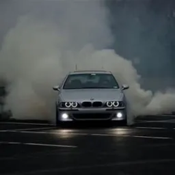 Srebrne BMW E39 z włączonymi ringami, unoszące się w chmurze dymu na asfaltowym parkingu. Dynamiczne ujęcie z przodu, podkreślające tuning wizualny.