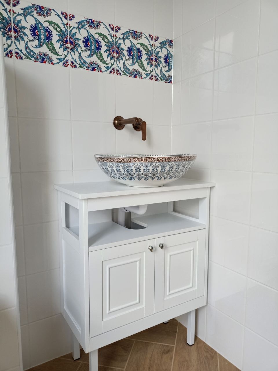 Biała szafka łazienkowa z umywalką w stylu orientalnym, dekoracyjne płytki na ścianie, brązowa armatura. Wykończona łazienka z dbałością o detale.