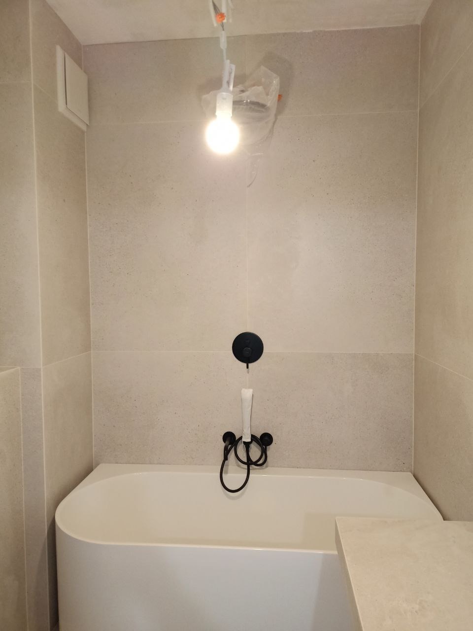 Minimalistyczna łazienka z wanną, czarną armaturą i beżowymi płytkami. Wisząca żarówka tymczasowa. Ujęcie z przodu, podkreślające nowoczesny design i stonowaną kolorystykę.