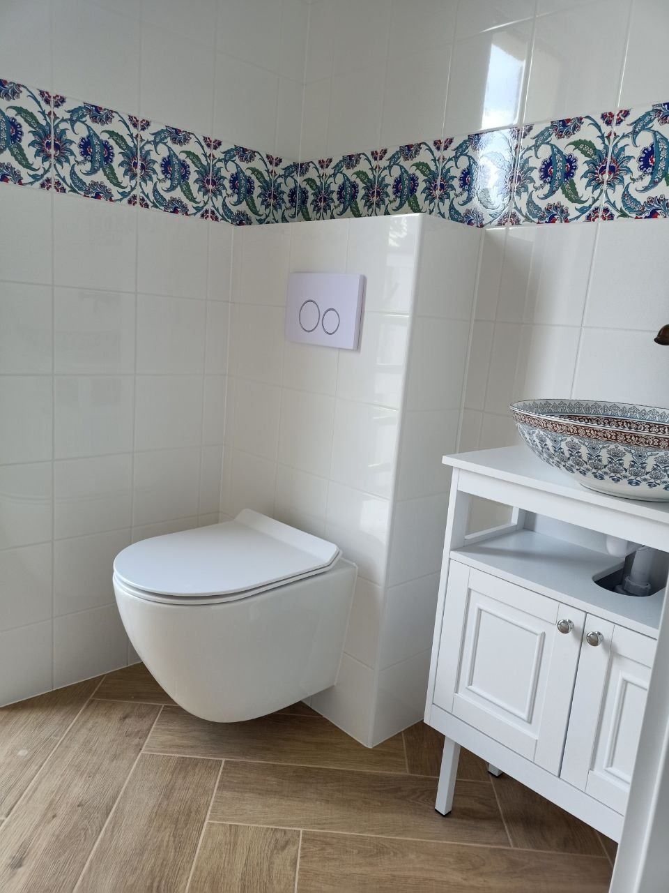 Nowoczesna łazienka z podwieszaną toaletą, umywalką w stylu marokańskim na białej szafce i dekoracyjnym pasem płytek nad białymi kafelkami. Drewnopodobna podłoga.