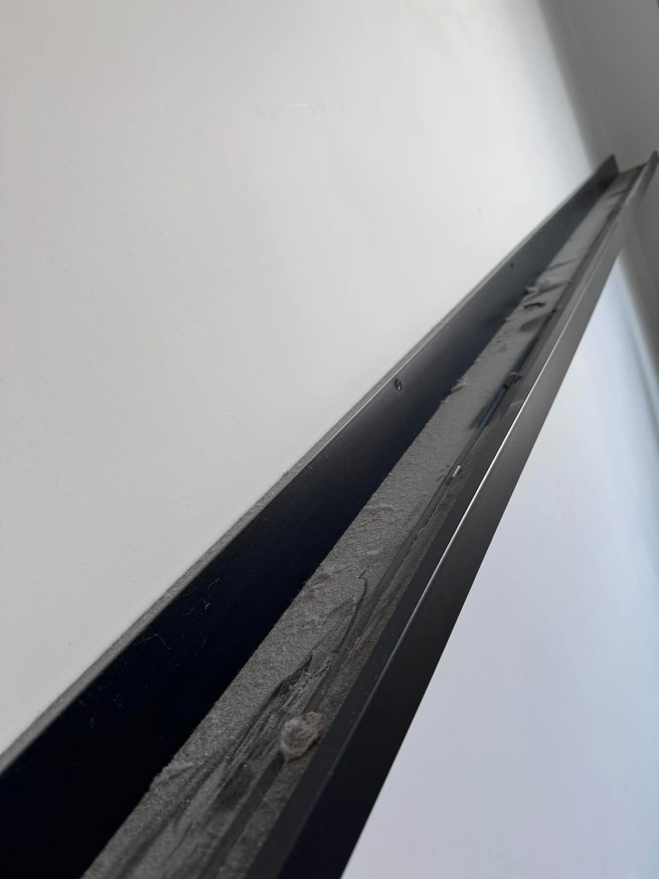 Zabrudzona listwa przysufitowa z kurzem i pajęczynami na tle białej ściany, widok z dołu pod kątem, ukazujący zaniedbanie i potrzebę sprzątania.