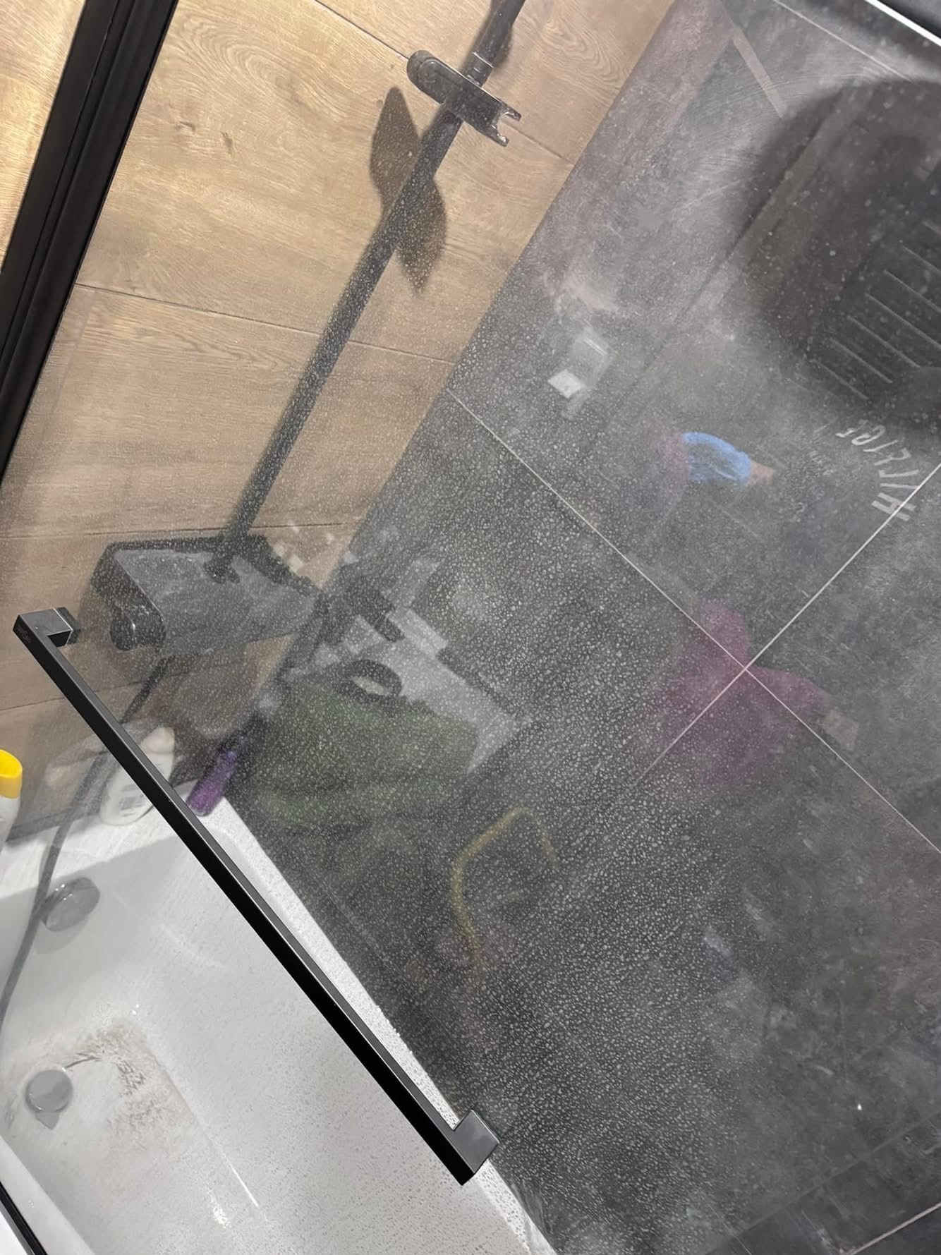 Zabrudzona kabina prysznicowa z widocznymi śladami osadu i zacieków na szklanych powierzchniach oraz ciemnych płytkach. Widoczny fragment baterii i akcesoriów łazienkowych.
