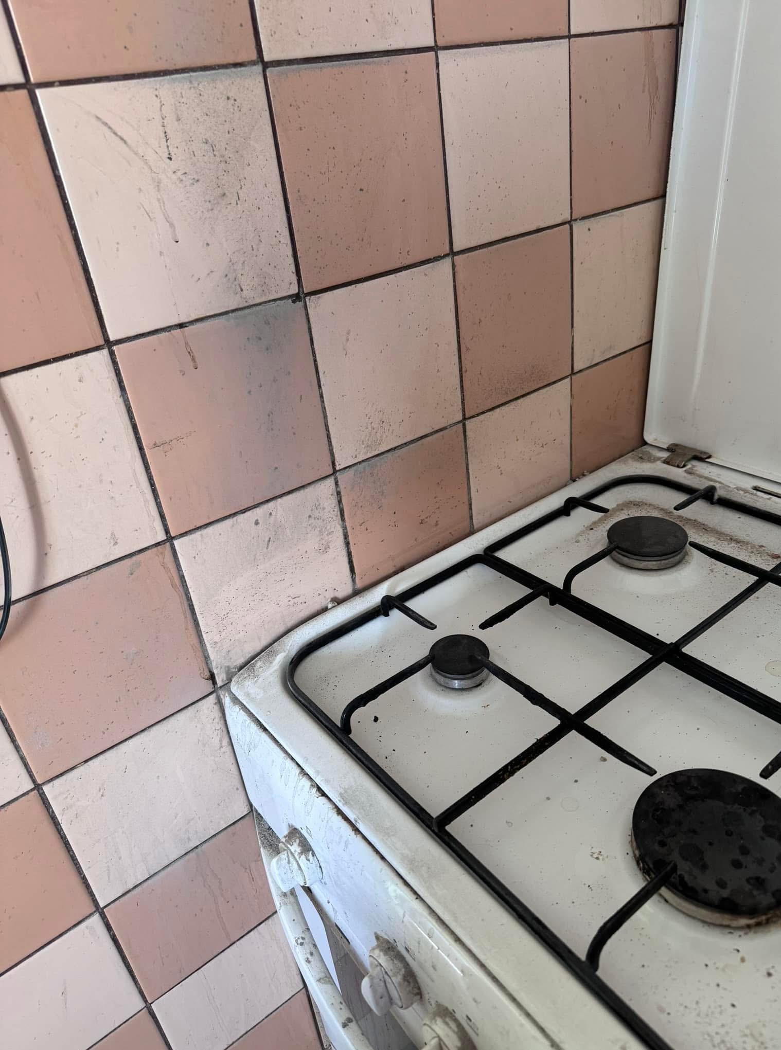 Zabrudzona, biała kuchenka gazowa z czarnymi rusztami i różowe, brudne kafelki na ścianie. Widoczne ślady tłuszczu i zabrudzeń. Potrzeba gruntownego czyszczenia.