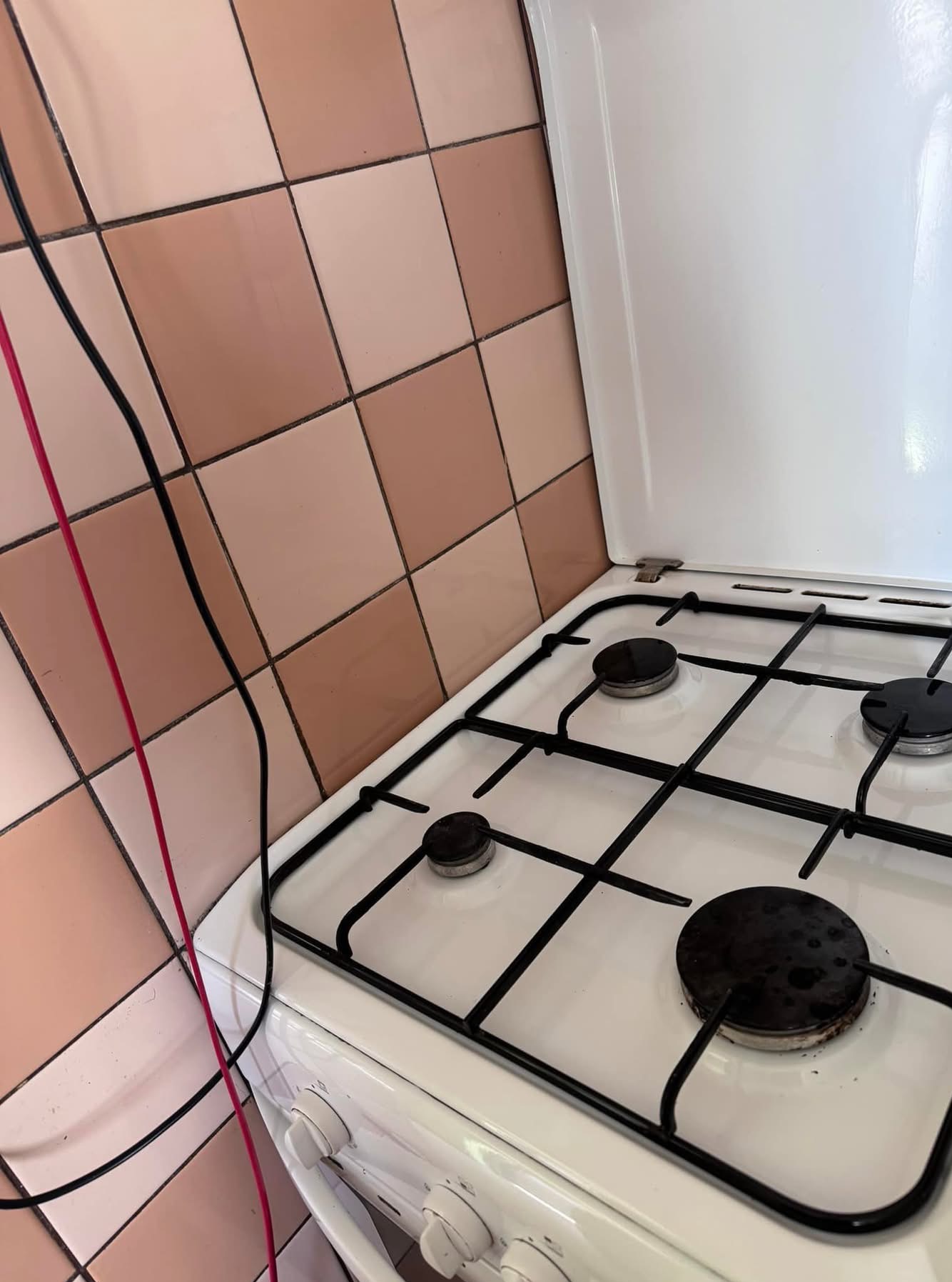 Biała kuchenka gazowa z czterema palnikami i brudnymi rusztami na tle różowych, kwadratowych płytek ściennych. Widoczne przewody elektryczne po lewej stronie.