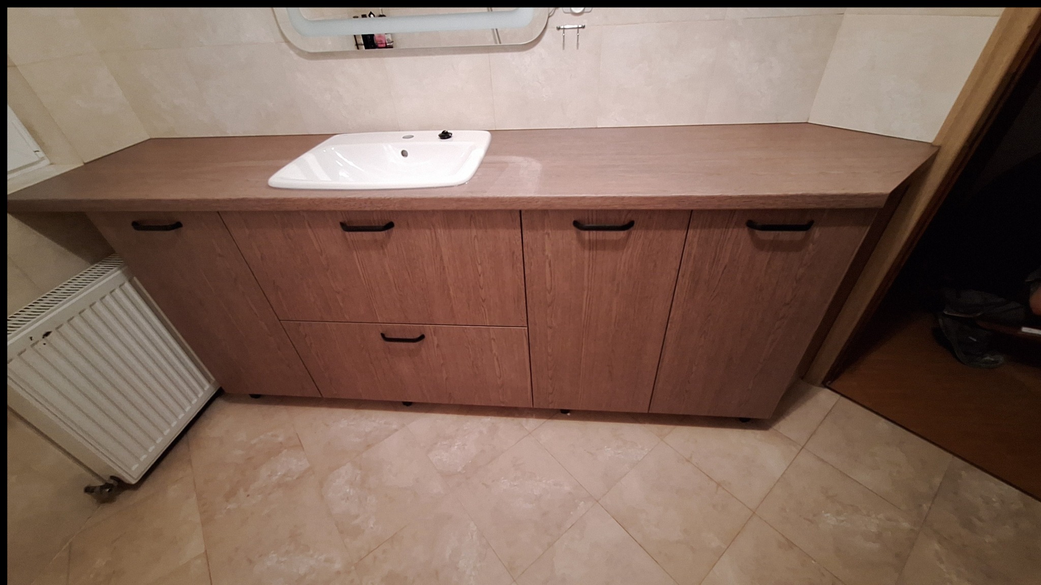 Jasna łazienka z szafką pod umywalką o drewnianym dekorze i czarnymi uchwytami, widoczny grzejnik i fragment otwartych drzwi.