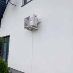 Biała jednostka zewnętrzna klimatyzacji Haier zamontowana na ścianie domu, z widoczną instalacją odprowadzającą skropliny. Nowoczesny design, czysty montaż.