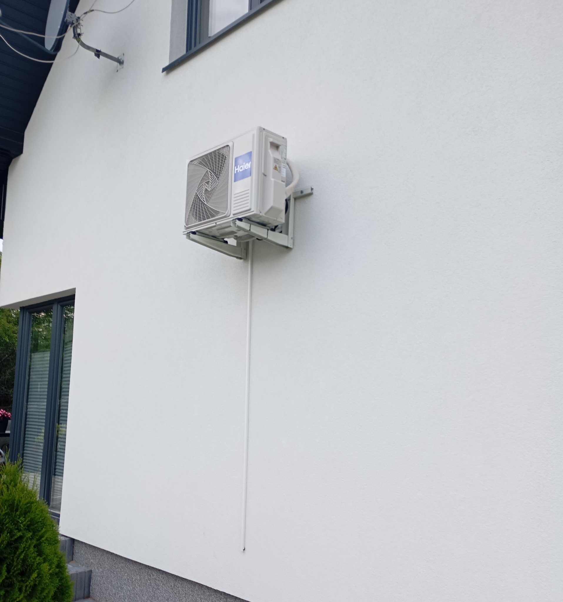 Biała jednostka zewnętrzna klimatyzacji Haier zamontowana na ścianie domu, z widoczną instalacją odprowadzającą skropliny. Nowoczesny design, czysty montaż.