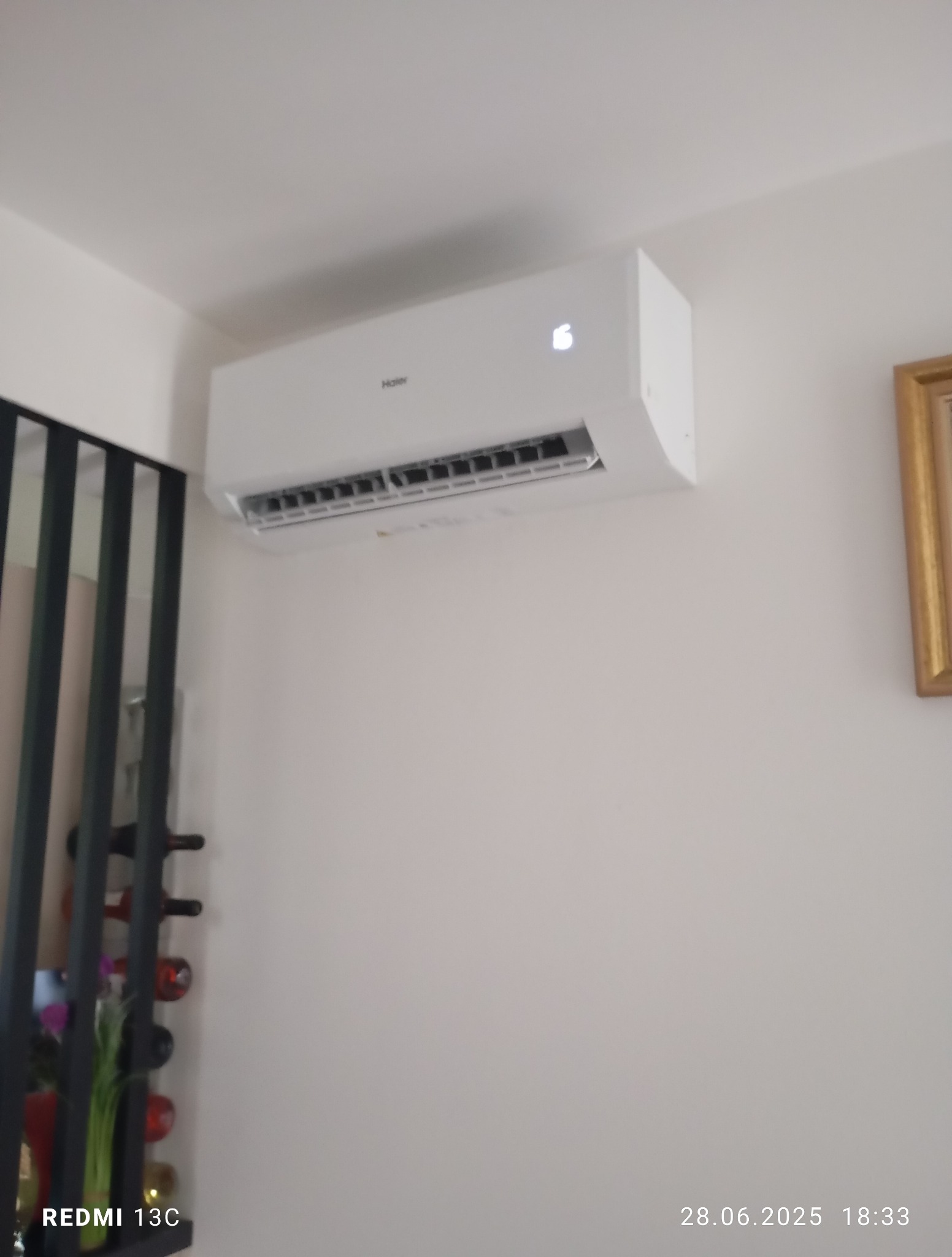 Biały klimatyzator Haier zamontowany na ścianie w pomieszczeniu, z widocznym wyświetlaczem temperatury i fragmentem dekoracji ściennej po lewej stronie.