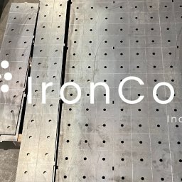 IronCore Aleksander Głowacki - Metalowe płyty z otworami ułożone na podłodze, gotowe elementy po obróbce, z widocznym logo firmy nałożonym na zdjęcie.