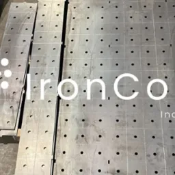 Metalowe płyty z otworami ułożone na podłodze, gotowe elementy po obróbce, z widocznym logo firmy nałożonym na zdjęcie.