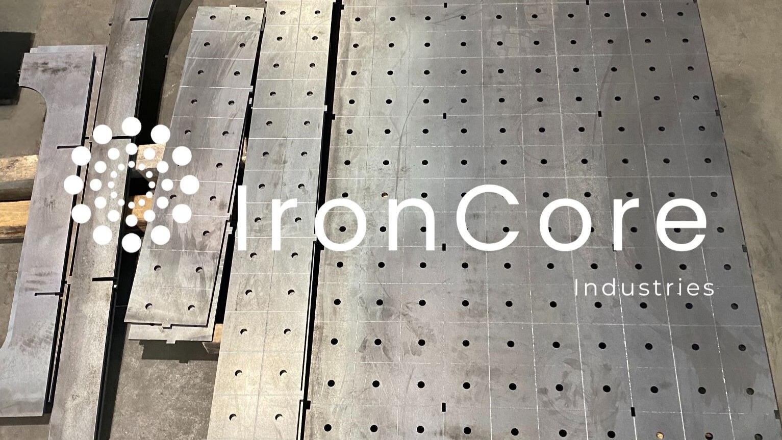 Metalowe płyty z otworami ułożone na podłodze, gotowe elementy po obróbce, z widocznym logo firmy nałożonym na zdjęcie.