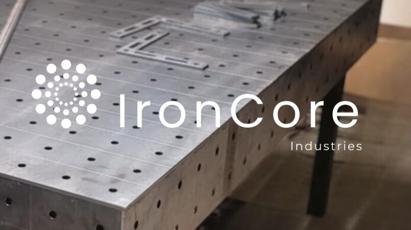 Stół spawalniczy z otworami, na nim metalowe elementy. Widoczne logo firmy IronCore Industries. Stół o szarej, metalicznej barwie, z równomiernie rozmieszczonymi otworami.