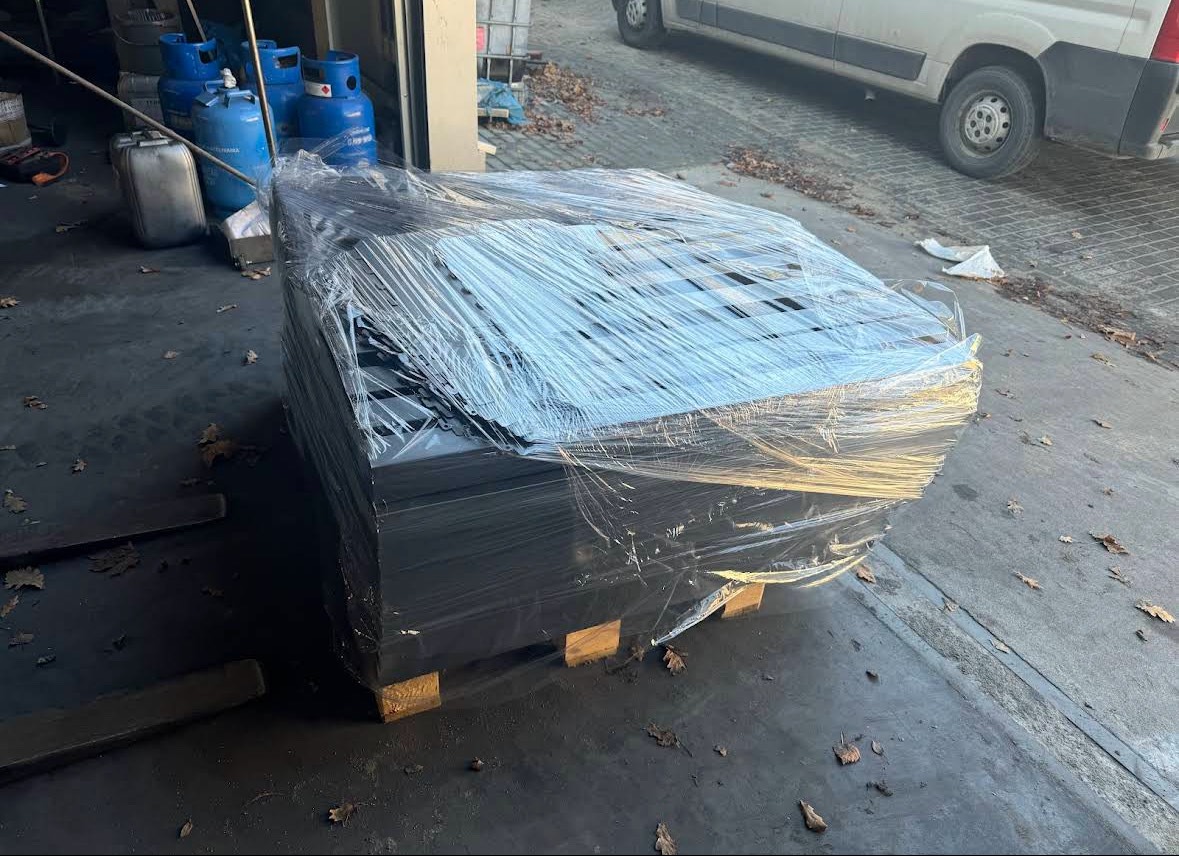 Czarne, perforowane blachy na palecie, owinięte folią stretch, czekają na transport. W tle widoczne butle gazowe i samochód dostawczy.