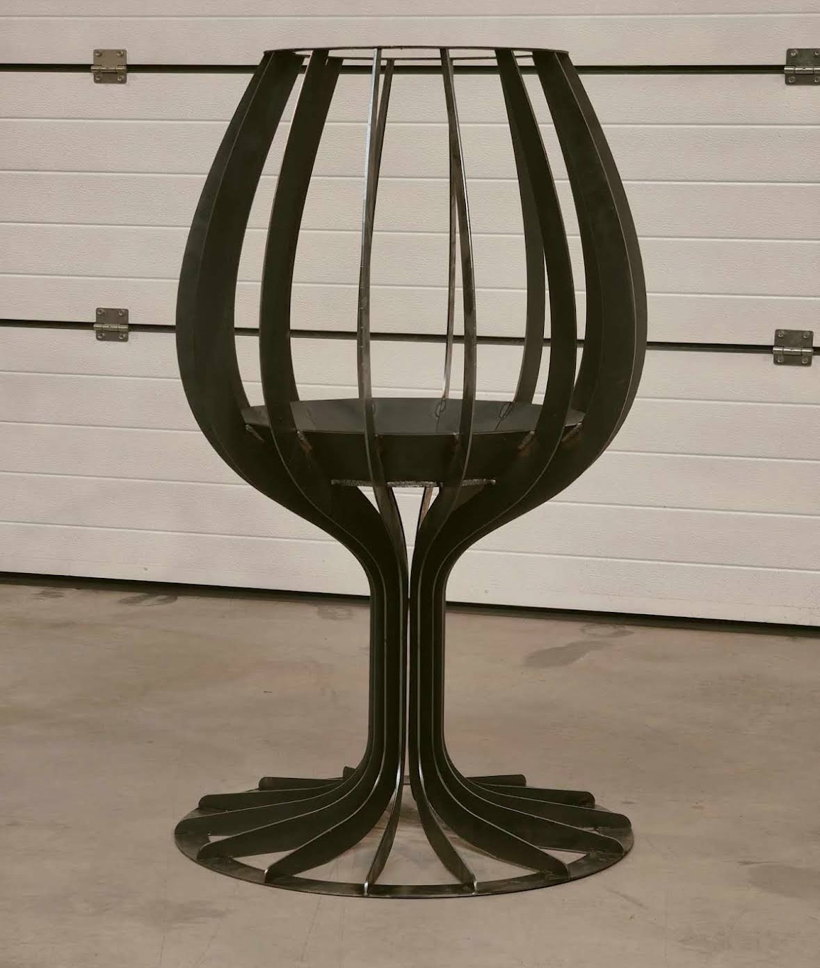 Metalowa konstrukcja przypominająca kielich, wykonana ze spawanych elementów, na tle białej bramy garażowej. Nowoczesny, minimalistyczny design.