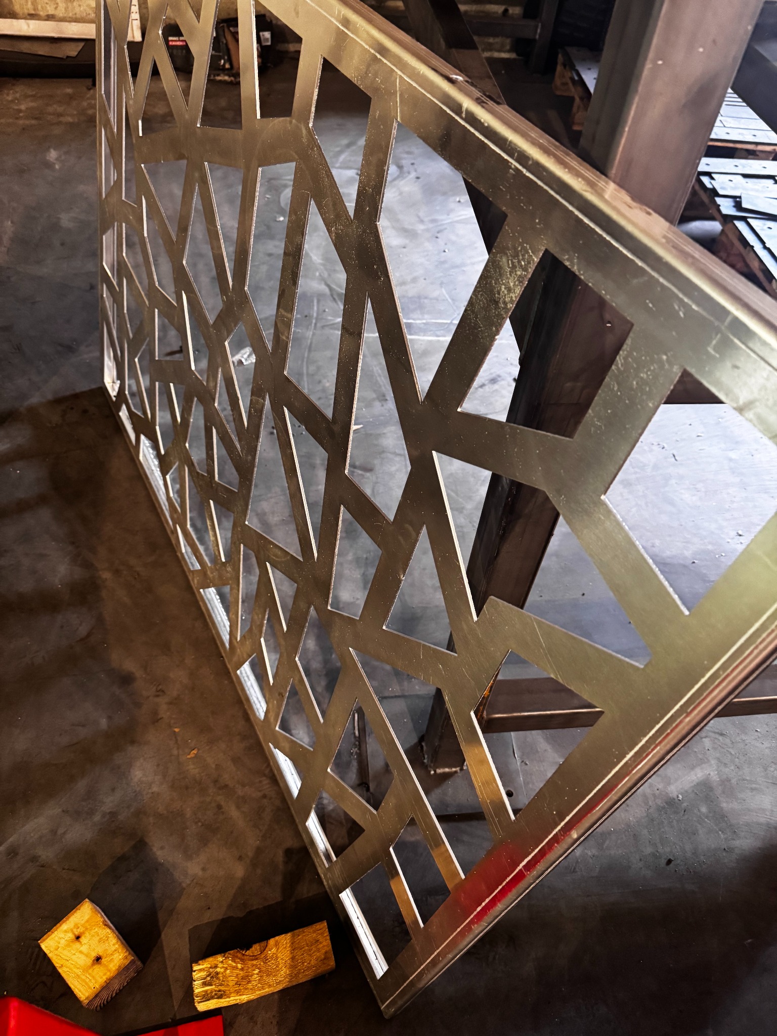 Metalowa płyta z wyciętym, geometrycznym wzorem, leżąca na podłodze warsztatu. Widoczne drewniane klocki i metalowe elementy konstrukcyjne w tle.