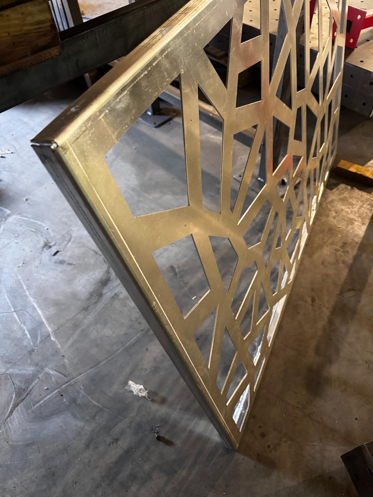 Metalowa konstrukcja z wyciętym wzorem geometrycznym, surowy wygląd, widoczne ślady obróbki, ustawiona ukośnie na betonowej posadzce.