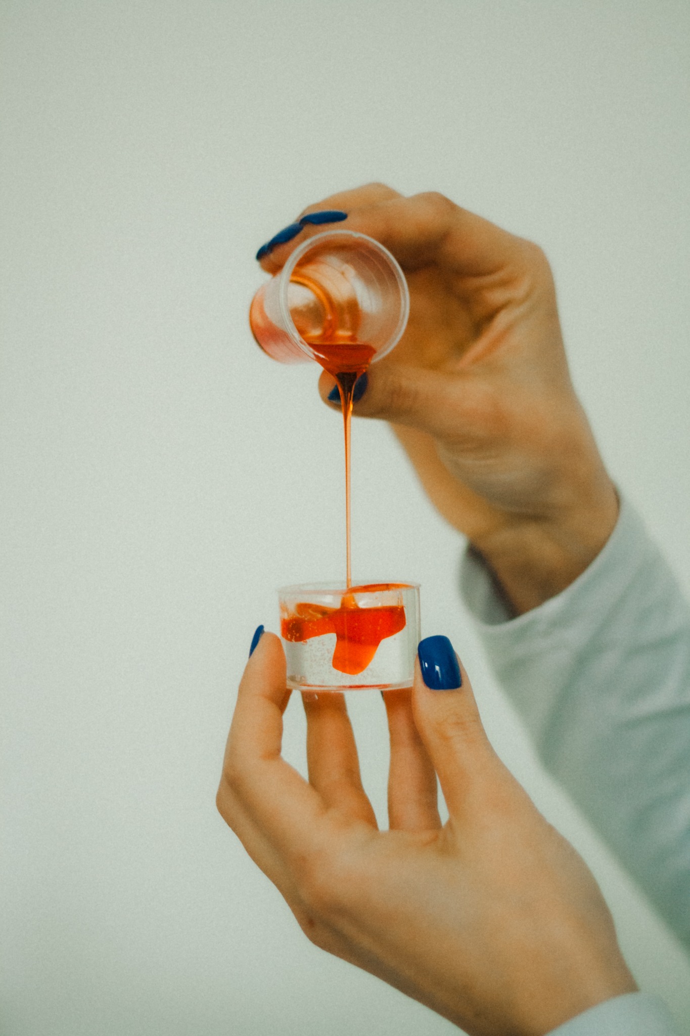 Dwie dłonie z granatowym manicure przelewają pomarańczowy płyn z jednego plastikowego kubka do drugiego na tle jasnej ściany.