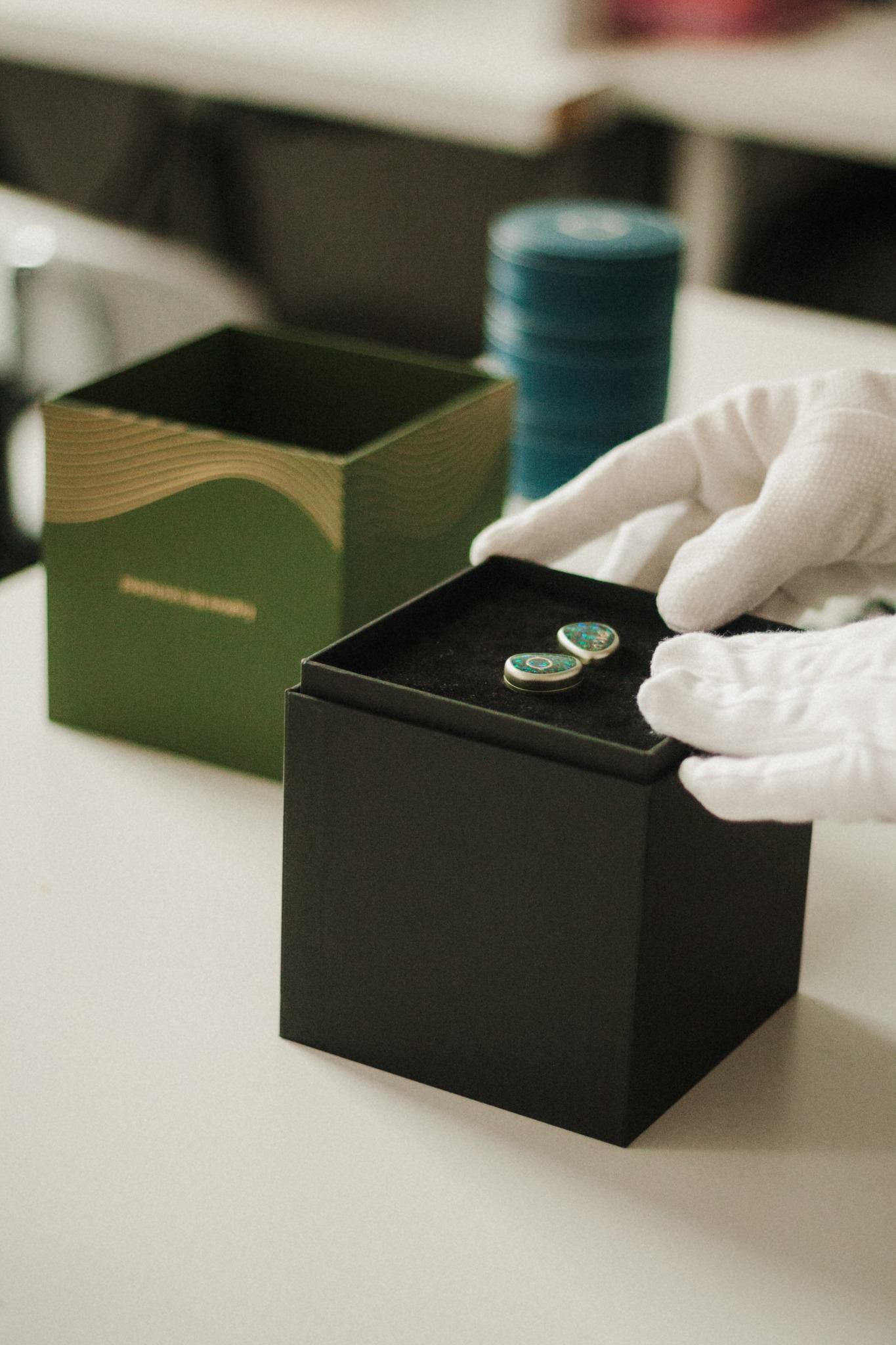 Dłonie w białych rękawiczkach układają parę kolczyków z turkusowymi detalami w czarnym pudełku na białym stole. W tle zielone pudełko z logo.
