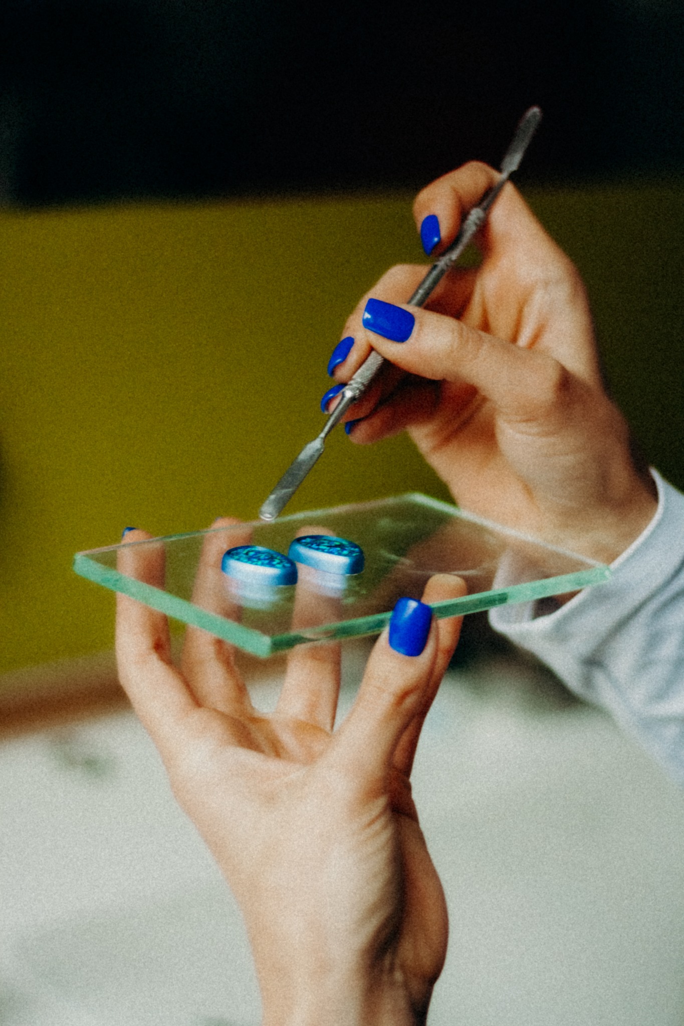 Dłonie z niebieskim manicure mieszają metalową szpatułką pigmenty na szklanej palecie, tworząc unikalne zdobienie paznokci w odcieniach błękitu i turkusu.