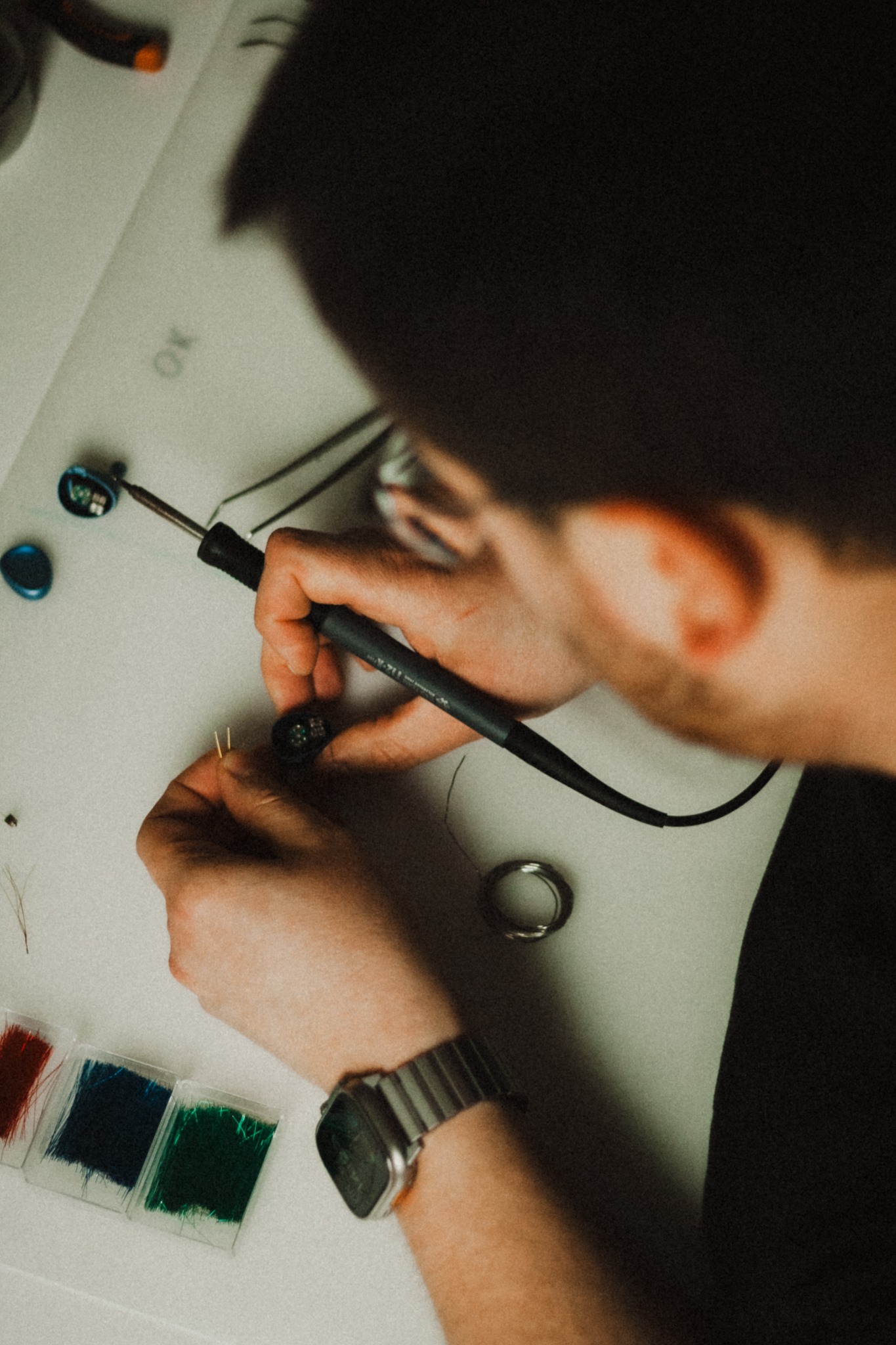 Osoba lutuje słuchawkę douszną na białym stole z narzędziami i kolorowymi elementami dekoracyjnymi. Zegarek na nadgarstku, widok z góry.