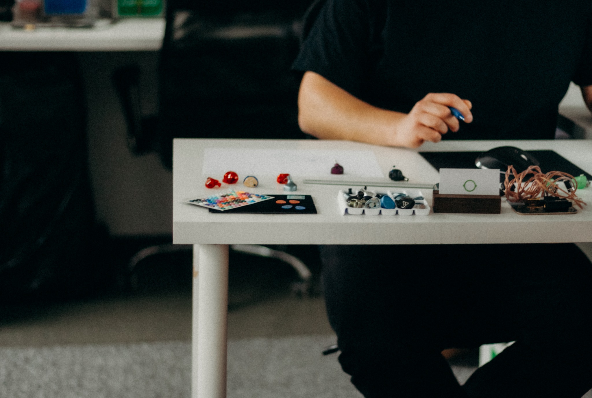 Osoba przy biurku projektuje słuchawki douszne, widoczne elementy i narzędzia do ich tworzenia. Ujęcie z bliska, stonowana kolorystyka.