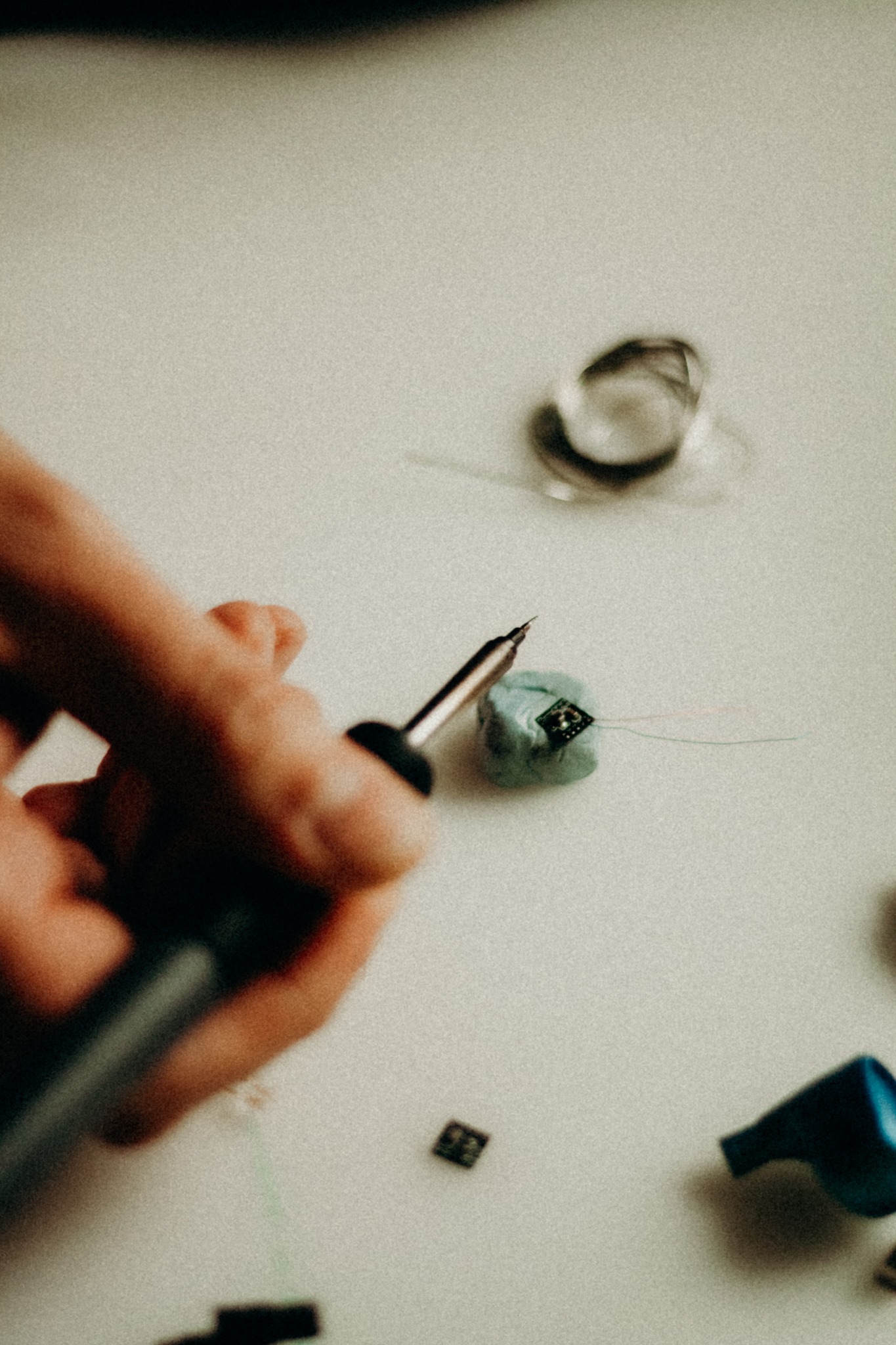 Ręka lutuje przewody do małego układu scalonego na jasnym tle. Wokół narzędzia, drut i elementy elektroniczne. Delikatne oświetlenie podkreśla precyzję pracy.