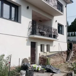 Biały dom z balkonami z kutą balustradą. W tle widoczne materiały budowlane i narzędzia, sugerujące prace wykończeniowe elewacji.