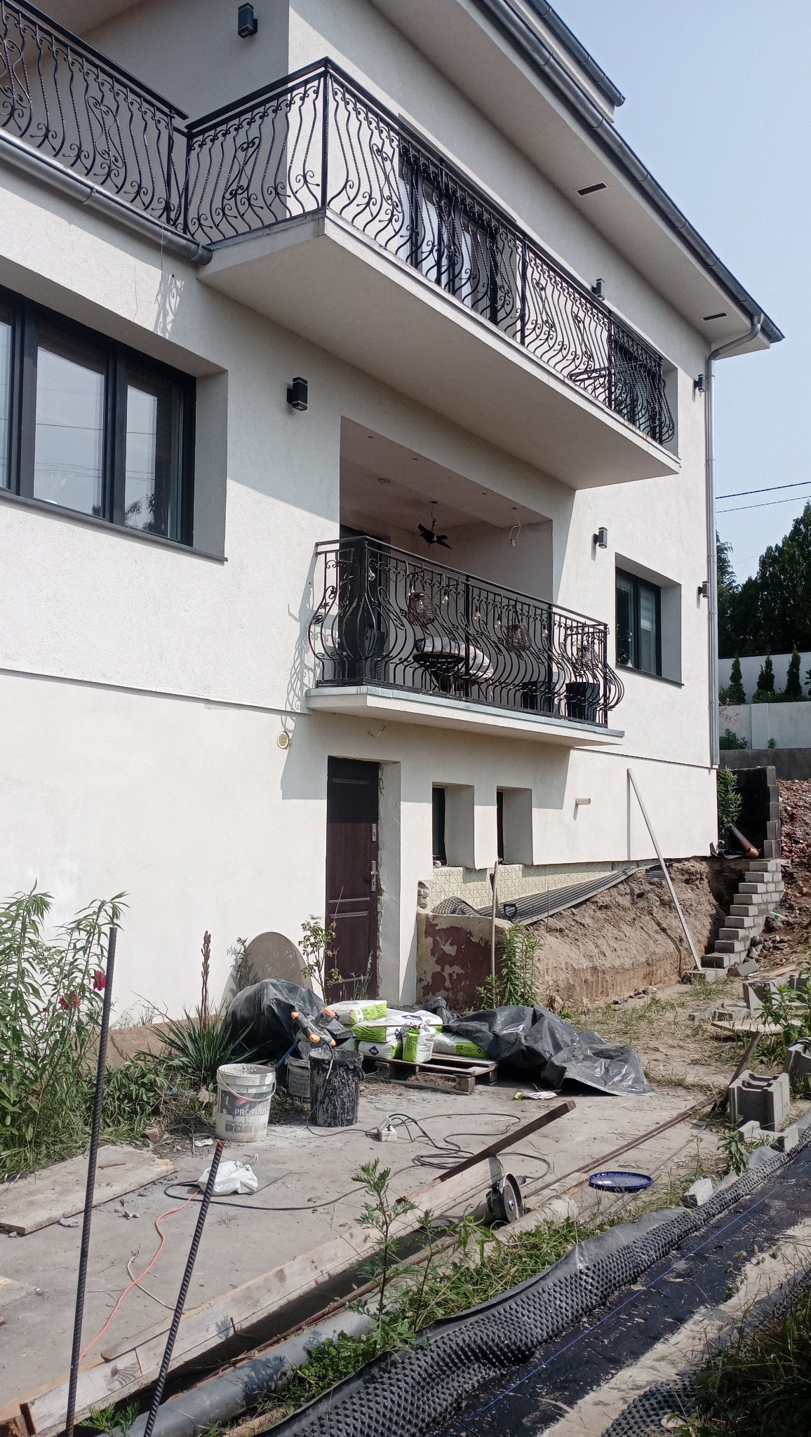 Biały dom z balkonami z kutą balustradą. W tle widoczne materiały budowlane i narzędzia, sugerujące prace wykończeniowe elewacji.