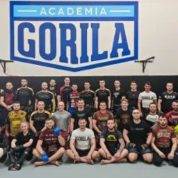 Grupowe zdjęcie z sali treningowej Academia Gorila Toruń. Mężczyźni i kobieta w strojach sportowych, część w rękawicach i ochraniaczach, siedzą na macie.