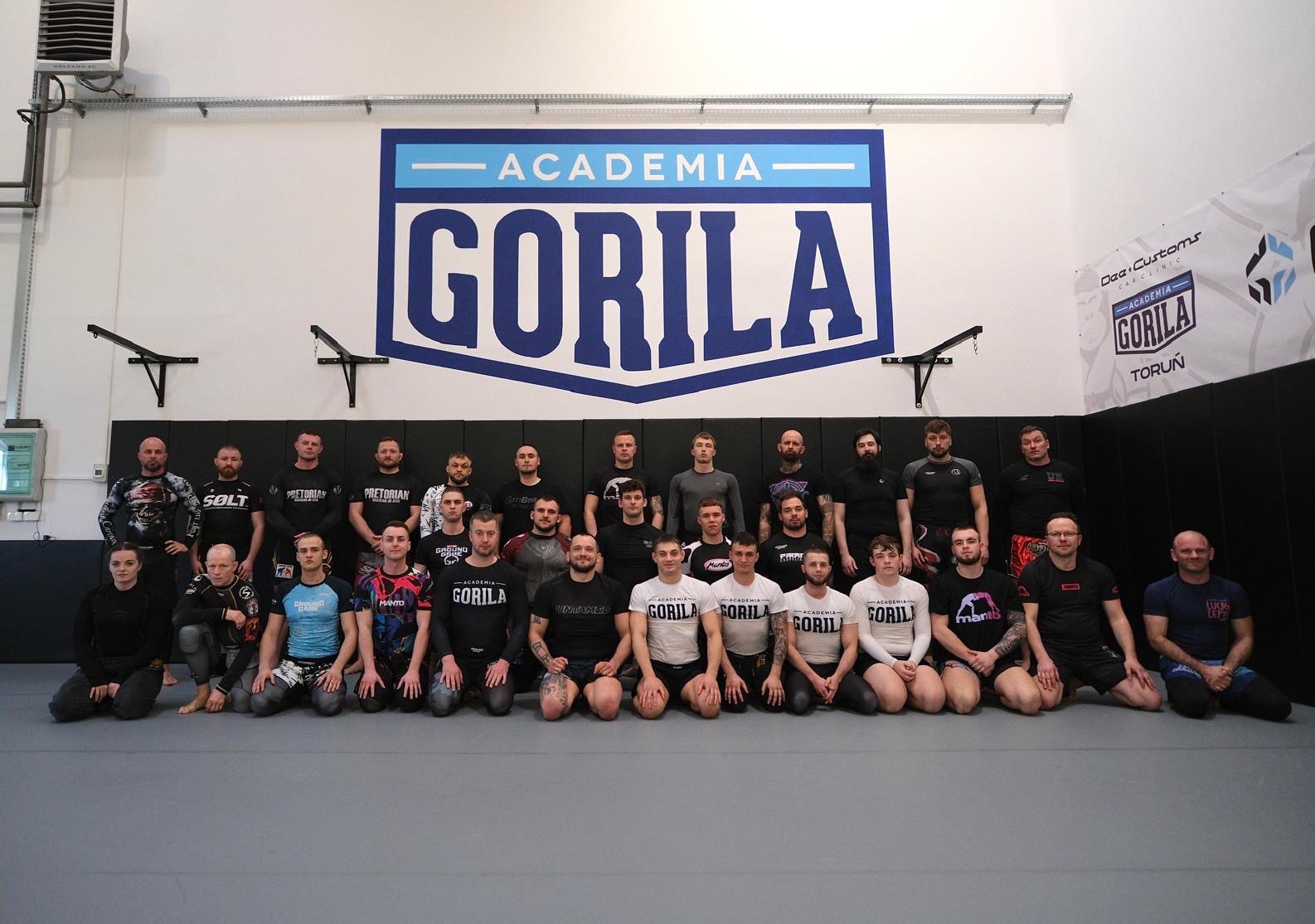 Grupowe zdjęcie uczestników Akademii Gorila Toruń na macie, z logo akademii w tle. Widoczni mężczyźni i kobiety w strojach sportowych, siedzący w rzędach.