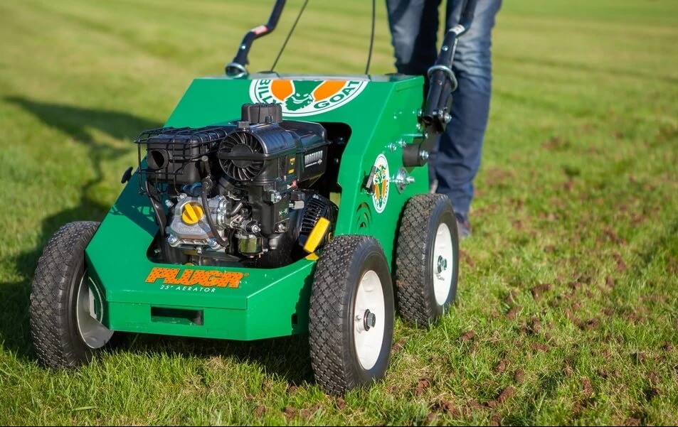 Aerator spalinowy Billy Goat na trawniku po aeracji, widoczne wyjęte rdzenie ziemi. Maszyna w kolorze zielonym z czarnym silnikiem i białymi kołami.