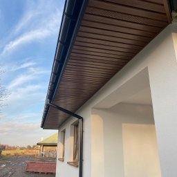 STREAM SP. Z O.O. SP.K. - Elewacja domu jednorodzinnego w jasnym kolorze, z drewnianą podbitką i orynnowaniem. Widok z dołu, na tle błękitnego nieba.